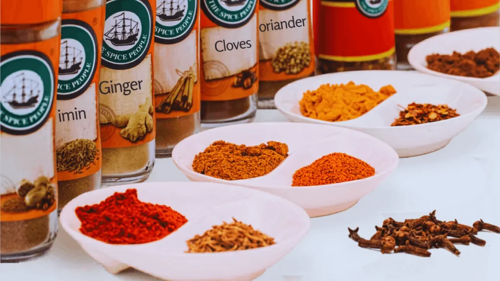 Various whole and ground spices in jars and white bowls including cumin, ginger, cloves, coriander, and paprika displayed for homemade BBQ spice rub recipe